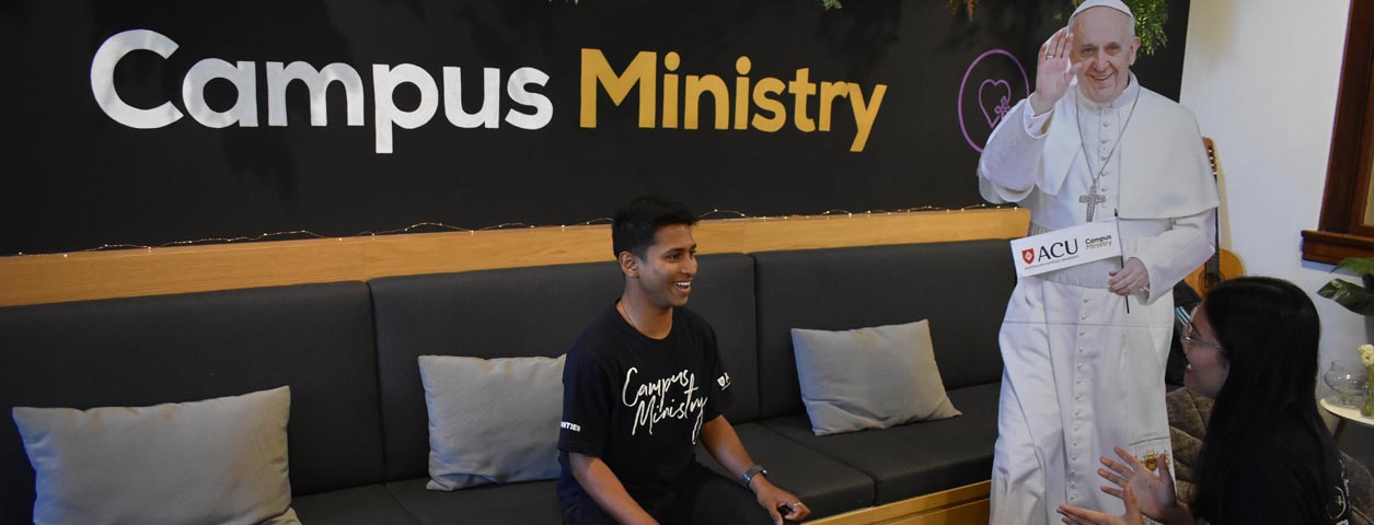 Two students sitting in the Campus Ministry Office, next to a life-size cut-out of the Pope.
